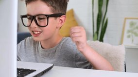 Happy young caucasian boy wearing glasses have a video call using a computer laptop while sitting at the table at home, smiling child waving his hands up and talks about his success