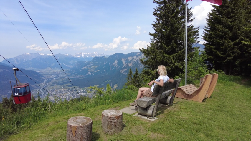 Woman relaxing on top of Brambruesch location in Switzerland. Swiss cable car of Chur or Coira with Swiss flag. Chur skyline in Grisons canton. Red cable car cabin from Chur to Kanzeli and Brambruesch