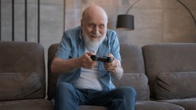 Portrait of an elderly man playing video game with wireless controller with joy of victory. Gray-haired pensioner, sitting on couch, emotionally plays on console, loses game, emotions of defeat.