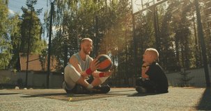 Sports happy family. Male basketball player spinning a ball on finger sitting with boy on basketball court