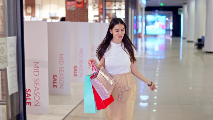 Attractive young brunette woman in trendy clothes jumping in excitement and being satisfied with her new purchases during shopping day in mall, carrying many colorful paper bags in hand Royalty-Free Stock Footage #1075469138
