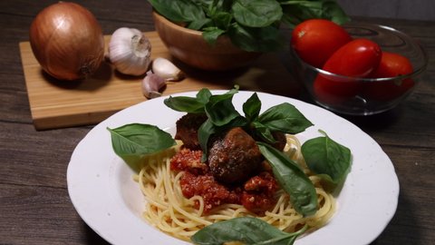 Sprinkle drop garnishing basil on cheese powder on meatball sauce cooked spaghetti white plate with ingredients roma tomato glass bowl onion garlic wooden board recipe on table marble wall 