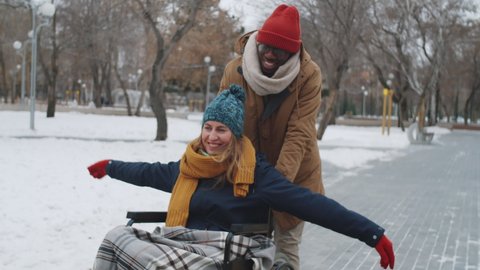Tilt up shot of young carefree woman smiling, chatting and holding arms outstretched while riding wheelchair with assistance of black male friend outdoors on winter day