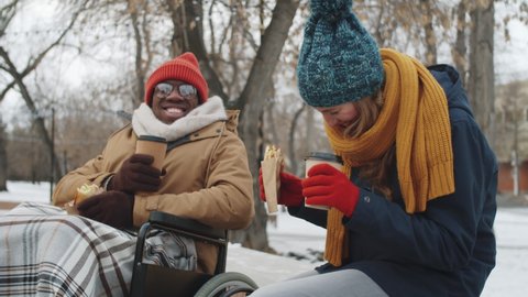 Young Afro-American man on wheelchair and Caucasian woman sitting outdoors on winter day, eating street food, drinking to go coffee, chatting and laughing