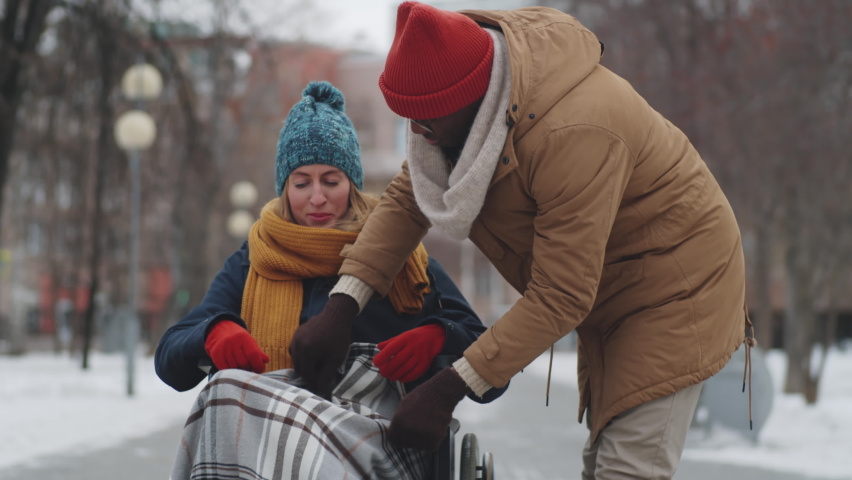 Afro-American man fixing blanket and pushing young cheerful woman on wheelchair while walking in park on winter day