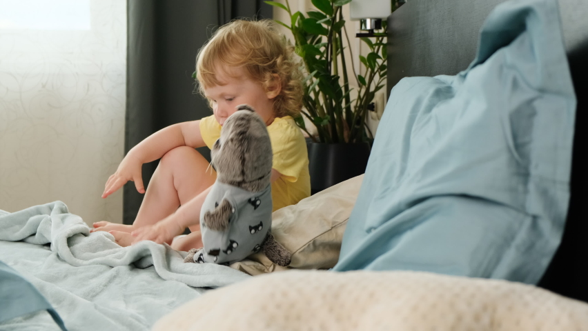 small beautiful child girl with blue eyes in yellow T-shirt prepares for sleep and goes to bed in parents bedroom and covers herself with blanket. Time for naps, day outside.
