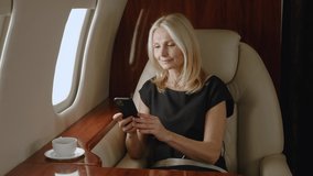 Mature rich woman using smartphone for video calling during luxury private jet travel. Gray haired beautiful senior woman having video conference on airplane