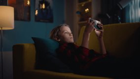 Excited Young Boy Lying on a Couch and Playing Video Games on Smartphone in Cozy Dark Room at Home. Happy Successful Teenager Beating Players and Winning at the Online Competition.