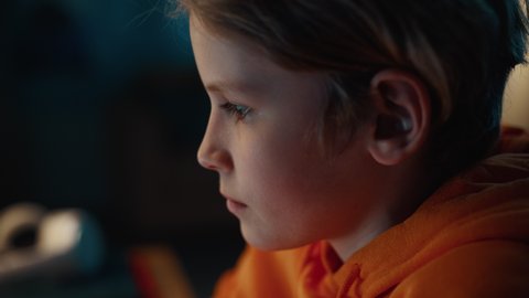 Portrait of a Cute Young Teenage Boy Being Concentrated on a Computer Screen in a Dark Cozy Room at Home. He's Wearing an Orange Hoodie. Curious Authentic Preteen Childhood and School Time Concept.
