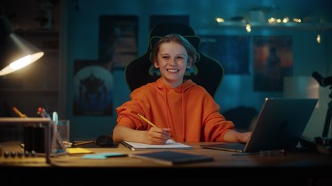 Portrait of a Smart Young Boy in Orange Sweatshirt Doing Homework on a Laptop Computer in Cozy Dark Room at Home. Teenager is Happy, Looks at the Camera and Smiles. Home Education Concept.