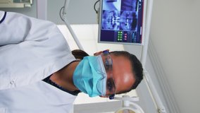 Vertical video: POV of patient in a dental clinic sitting on surgery chair checking affected mass. Dentistry team working in orthodontic office, lighting the lamp and examining person, close-up face