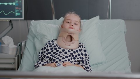 Portrait of hospitalized sick little child resting in bed wearing medical neck cervical collar during healthcare consultation in hospital ward. Disabled kid suffering painful accident