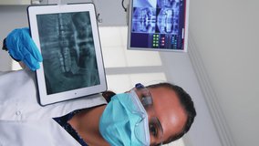 Vertical video: Dentist showing on tablet teeth x-ray reviewing it with patient. Doctor and nurse working together in modern stomatological clinic, explaining to old woman radiography of tooth using