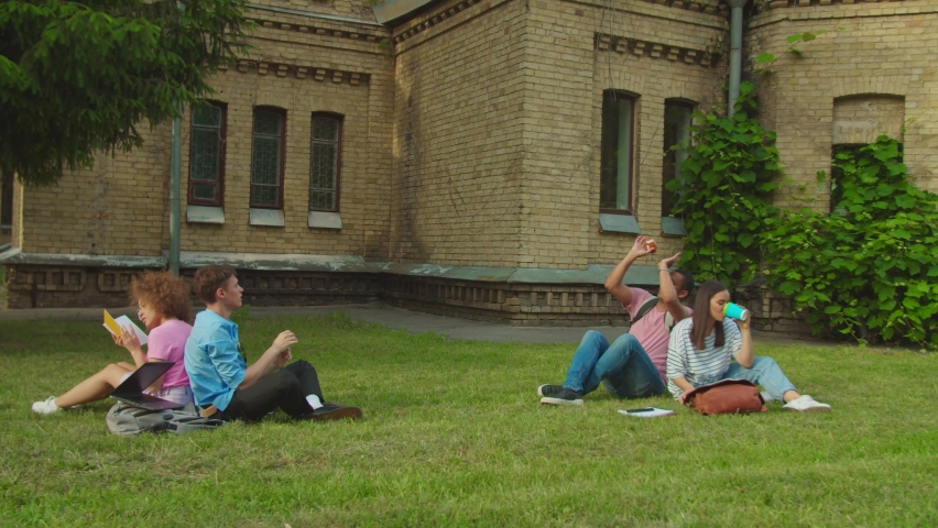 Group of mixed race students sitting on campus lawn outdoors, while two carefree boys throwing toy ball having fun, relaxing and two girls studying books drinking takeaway coffee outside