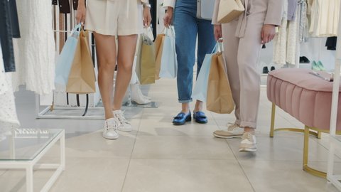 Low shot of female legs walking in store with shopping bags while freinds are buying clothing and shoes. Shelves with garments and footwear are visible in background