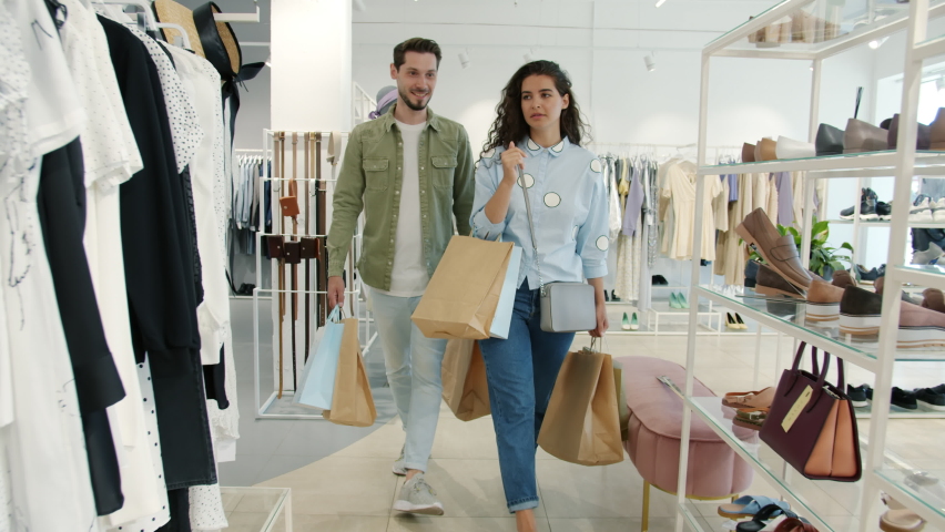 Cheerful man and woman walking in clothes store holding bags talking and smiling looking around at garments on hangers. Youth and consumerism concept. Royalty-Free Stock Footage #1075493126