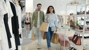 Cheerful man and woman walking in clothes store holding bags talking and smiling looking around at garments on hangers. Youth and consumerism concept.