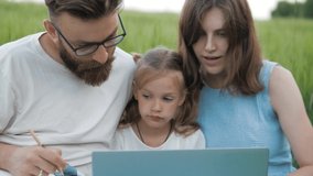 Cute family looking at laptop screen searching any information outdoors on a field.Mother, father and cute little daughter using laptop, making notes 