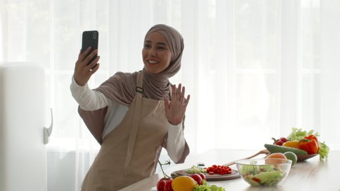 Food Blogging. Muslim Blogger Lady Making Video Recipe Cooking And Using Smartphone, Talking To Phone Camera And Showing Meal Ingredients In Modern Kitchen At Home, Wearing Hijab. Tracking Shot