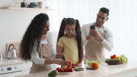 Modern Middle-Eastern Family Cooking Together, Arab Man Taking Photo On Smartphone While His Wife And Daughter Making Salad And Cutting Fresh Vegetables. Healthy Food And Nutrition
