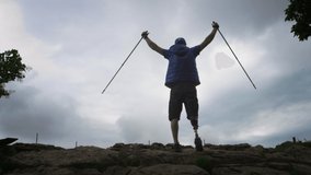 Male hiker with a prosthetic leg. Disabled man breaks through rocks, enjoying his time on a trip to the mountains, hiking, standing on a rock, raising his hands, holding Nordic walking sticks - Powered by Shutterstock - Get 15% off with code: PIKWIZARD15