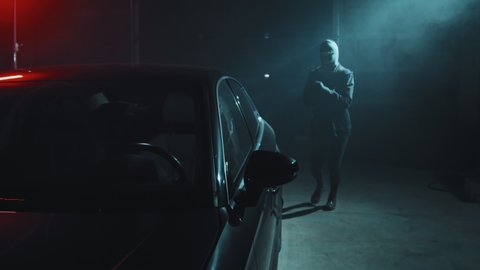 Slowmo tracking shot of masked burglar breaking into car parked in dark garage