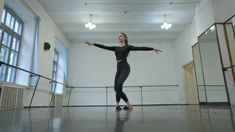 Wide shot of gorgeous confident slim ballerina spinning in dance studio indoors. Portrait of beautiful smiling Caucasian young woman rehearsing performance in slow motion. Art and grace concept