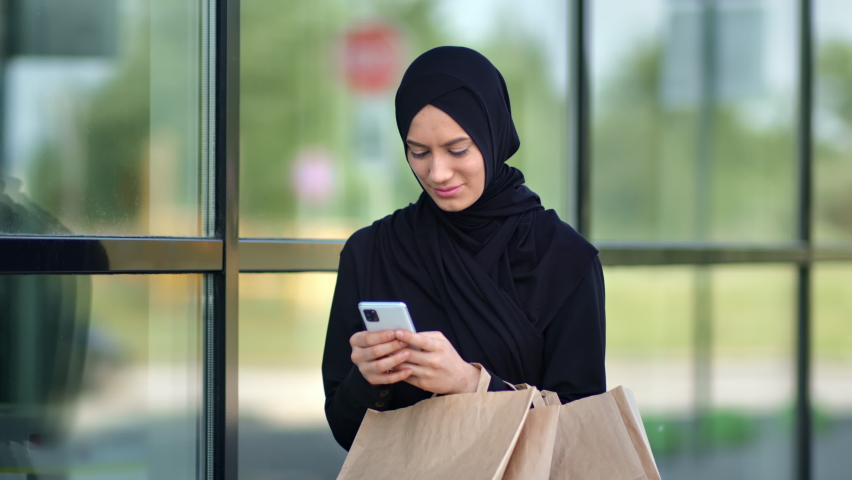 Smiling Muslim woman buyer chatting use smartphone holding shopping paper bag at glass building mall center outdoor. Happy shopaholic Islamic lady in black headscarf surfing internet on mobile phone  | Shutterstock HD Video #1075508087