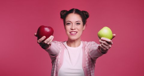 Beautiful young caucasian girl holds two apples, red and green in her hands and funny bites quickly each one in turn isolated on pibk background. Vegetarian healthy fruits. Problem of choice concept