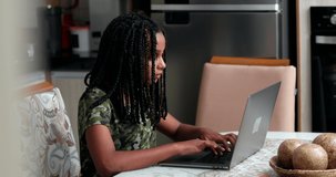 African child typing on laptop at home kitchen. Candid Black girl using computer