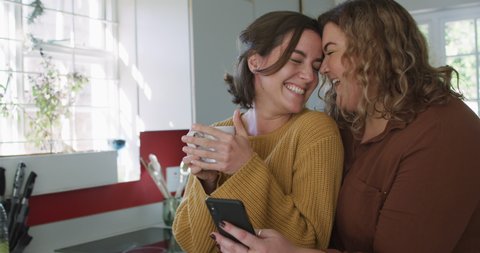 Happy caucasian lesbian couple embracing and using smartphone in kitchen. domestic life and leisure time.
