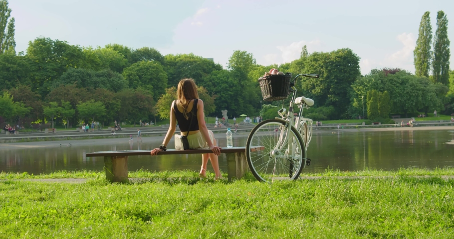 A beautiful girl is resting on a bench in the city park next to a white bicycle. Caucasian woman sitting on a wooden bench relaxing in front of the lake. Relaxing girl after biking on vacation. Royalty-Free Stock Footage #1075522226