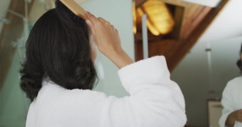 Mixed race woman wearing bathrobe looking at mirror and brushing her hair. domestic life, spending quality free time relaxing at home.