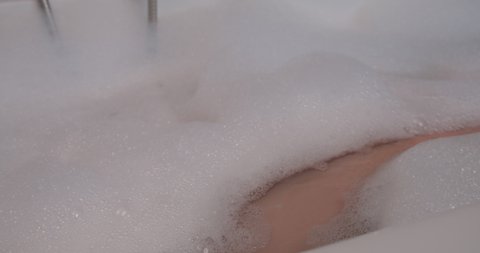 One leg sticks up out of a relaxing bubble bath and is cleaned with an exfoliating glove