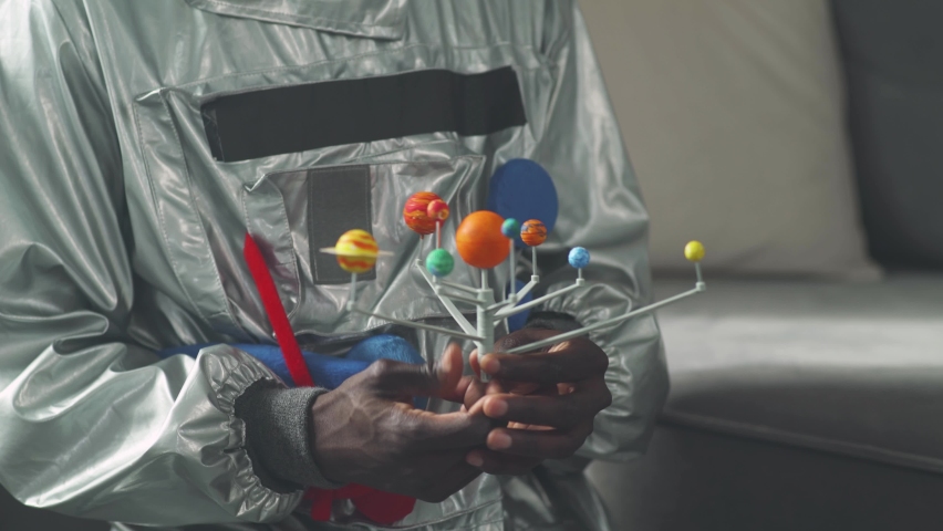 African man in an astronaut costume sitting on the floor in living room at home and playing with a toy model of the solar system, study of astronomy, man on self-isolation.