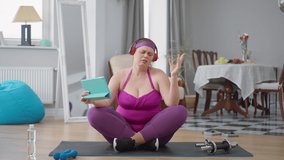 Confident plus-size Caucasian woman enjoying music in headphones sitting on exercise mat filming video blog. Obese young lady blogging training at home indoors. Confidence and self improvement