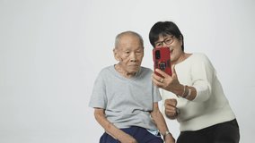 Senior Asian man doing a video call via mobile phone to his relative with his daughter. A 95 years old guy with a cataract eye problem learns how to take a selfie.