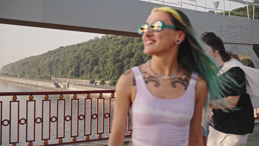 Active girl with bright hair and a tattoo is riding a skateboard across the bridge in the company of her friends, skateboarders. Youth culture, urban landscape.