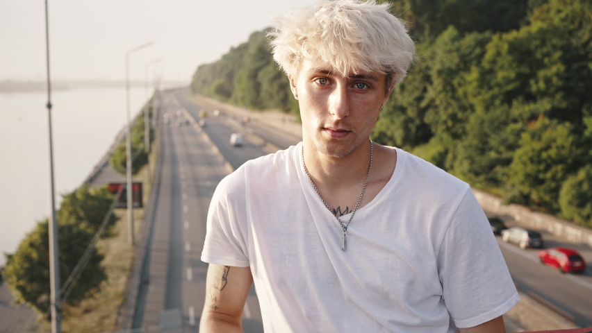 Close up portrait of blond guy on the bridge. A man with a bright appearance, dyed hair and tattoos looking at the camera.
