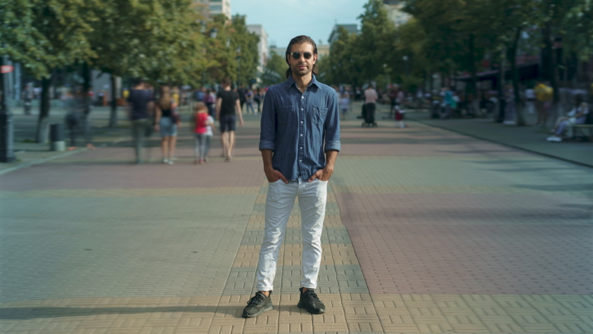 Zoom-in time lapse of attractive Middle Eastern man standing outside in crowded street with people rushing around. Summertime and youth concept.