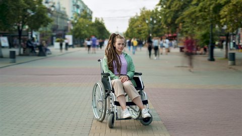 Zoom-in time lapse of attractive disabled girl with dreads hairdo sitting in wheelchair in busy street city while crowds of people walking around