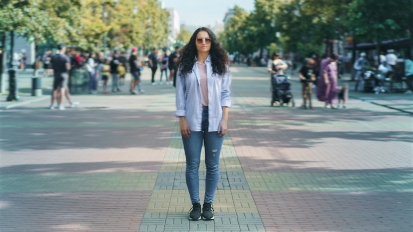 Zoom-in time lapse portrait of serious Asian girl standing in pedestrian street in modern city while youth are walking and having fun around. Millennials and society concept.