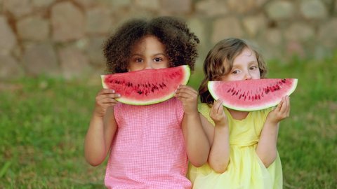 Funny Black girl and European girl are eating watermelon outdoors in the hot summer. Smiling children in light dresses enjoy healthy food outdoor