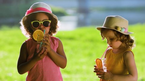 Portrait of a white and Black friends in a summer park. kids in summer dresses and a hats outdoors . Childrens suck from the straw a fresh juice