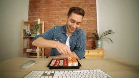 Front view of happy handsome young man eating sushi during watching or reading information from monitor screen, looking at camera. POV of bearded Caucasian male eating rolls watching movie at desk.