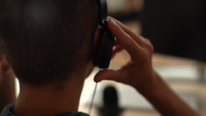 Close-up back view of unrecognizable blogger male putting headphones on head sitting at desk with microphone and computer monitor. Closeup rear view of young man puts headphones on head indoors. Royalty-Free Stock Footage #1075558163