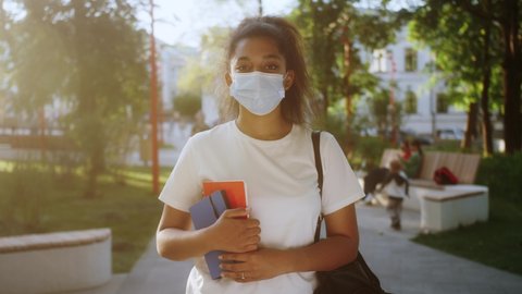 Happy mixed race teen girl school college student wearing medical mask, smiling and looking at camera. Portrait of student outdoor.