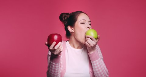 Beautiful young caucasian girl holds two apples, red and green in her hands and funny bites quickly each one in turn isolated on pibk background. Vegetarian healthy fruits. Problem of choice concept