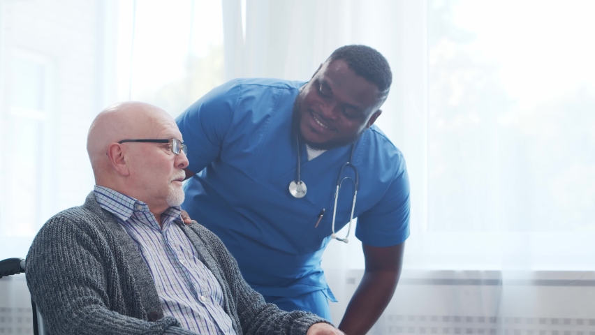 African-American caregiver and old disabled man in a wheelchair. Professional nurse and handicapped patient in a nursing home. Assistance, rehabilitation and health care. Royalty-Free Stock Footage #1075563722