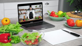 Close up laptop computer with man chef blogger in screen tells recipe dish teaches student stands on kitchen table near food ingredients and cutting board with knife, online video call culinary lesson